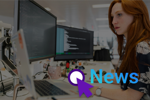 Sock Foto. Eine rothaarige junge Frau arbeitet an ihrem Platz mit Laptop und zwei Bildschirmen in einem Büro. Rechts unten steht das wort "News" in blau neben einem lila Cursor-Symbol.