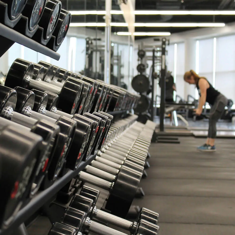 Rows of dumbbells on racks in a gym with a woman exercising in the background.