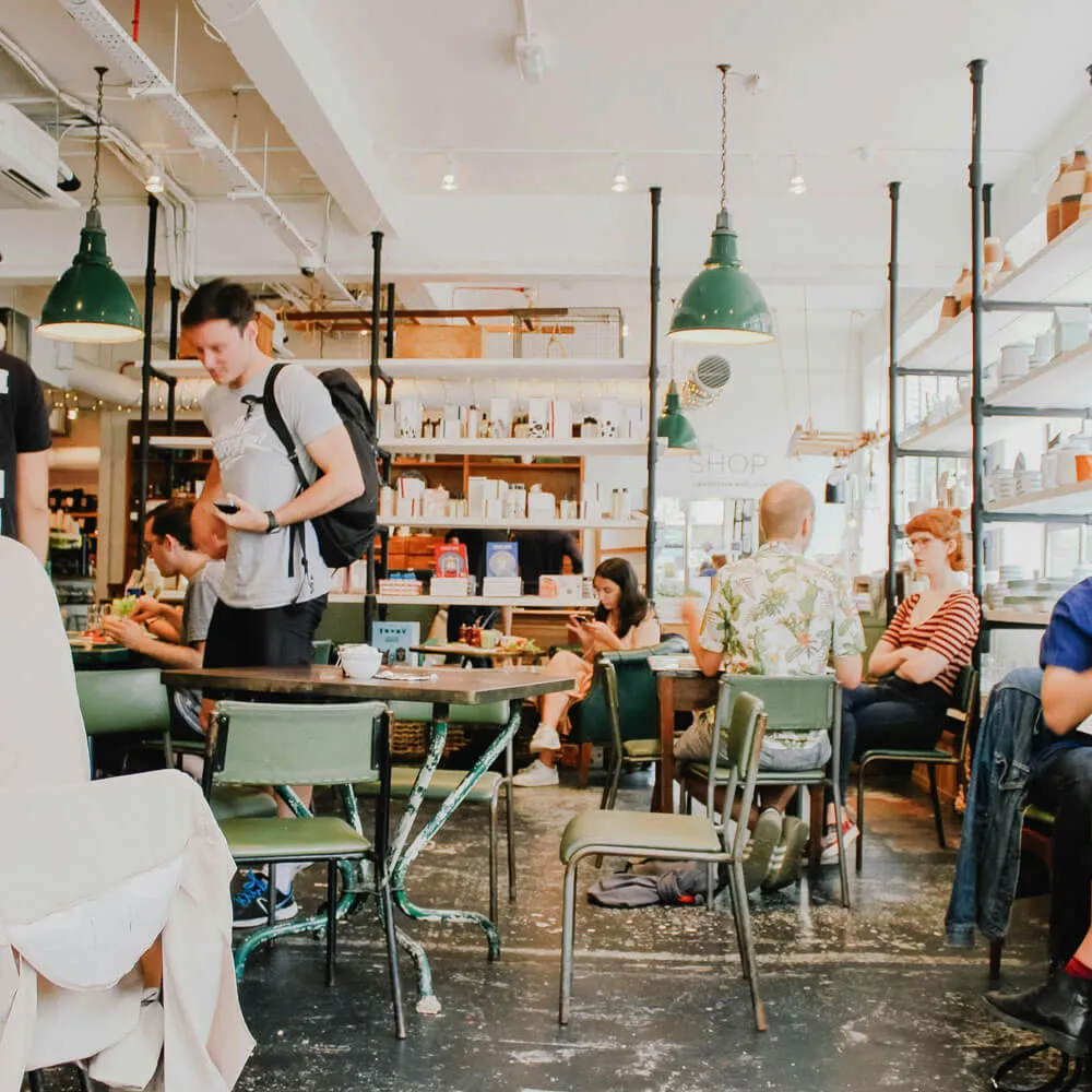 People sitting and standing in a cozy café with green chairs, hanging green lamps, and shelves of products.
