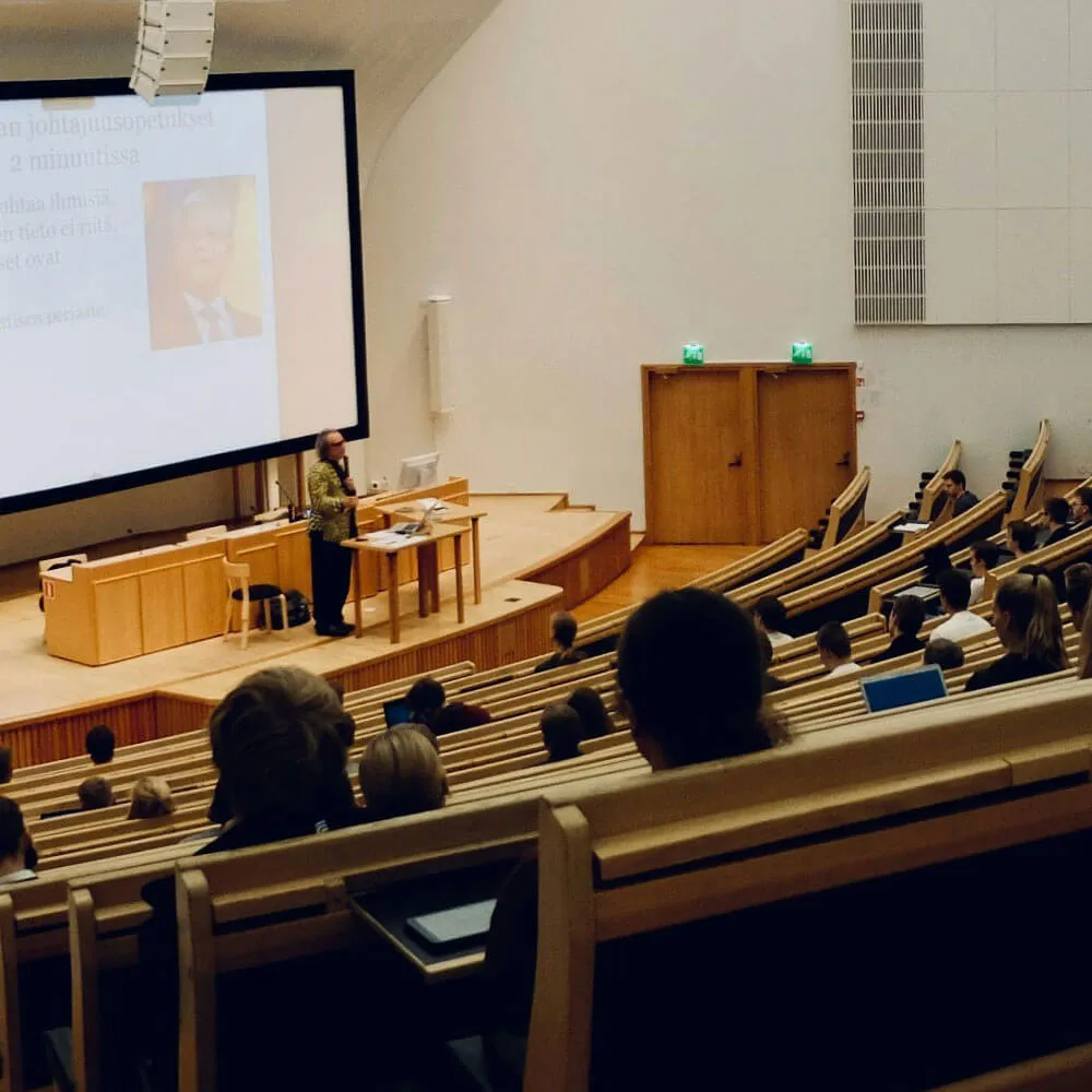 University lecture hall with a female professor presenting to seated students and a large projection screen.