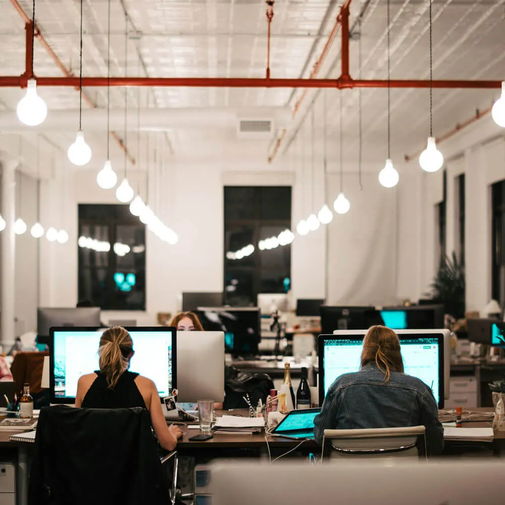 Two women working at computers in a modern office with hanging light bulbs and a spacious layout.