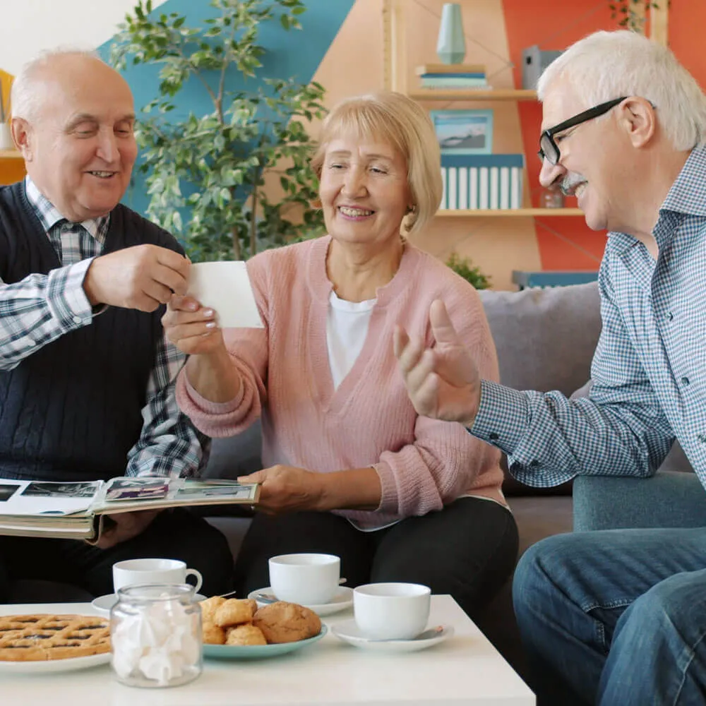 Three elderly people sitting on a couch, looking at a photo and smiling, with a table in front holding cups, cookies, and marshmallows.