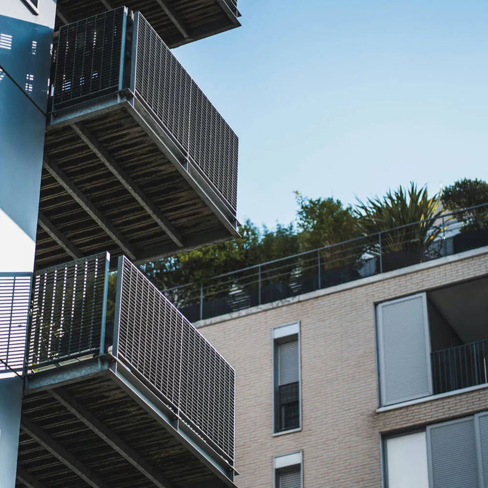 Close-up of metal balconies on a modern apartment building with greenery on the rooftop against a clear blue sky.