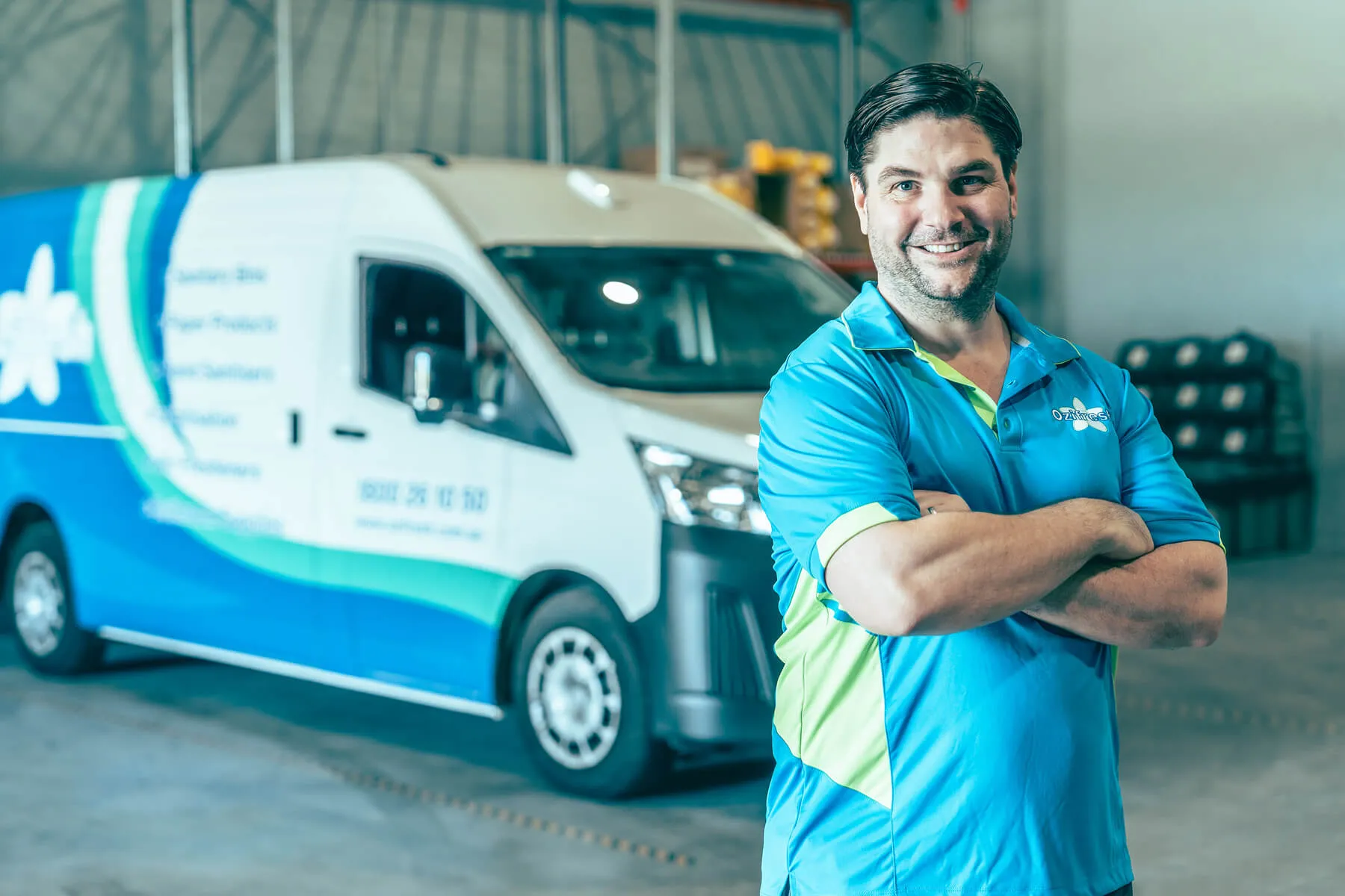 Smiling man with folded arms wearing a blue and green work shirt standing in a warehouse with a white and blue service van in the background.