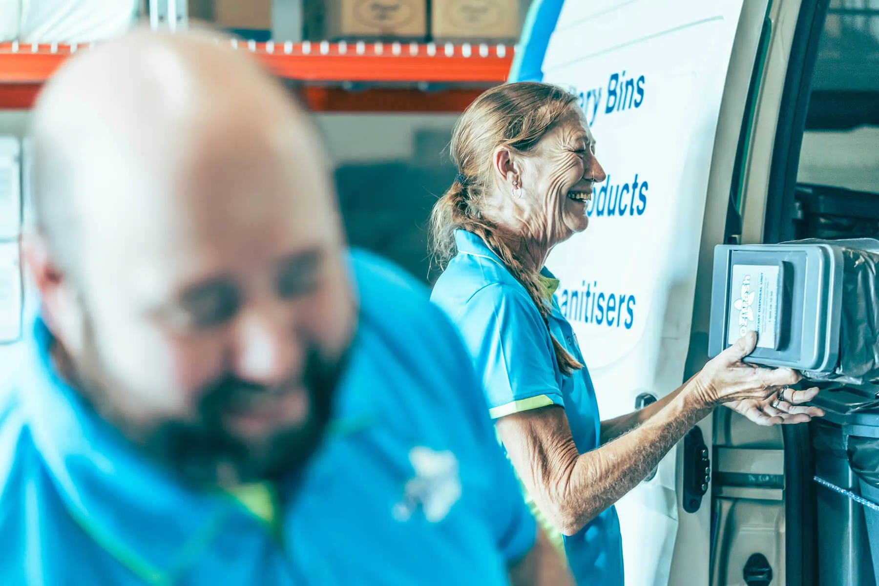 Two workers in blue uniforms smiling while handling equipment near a van with text about sanitary products and sanitizers.