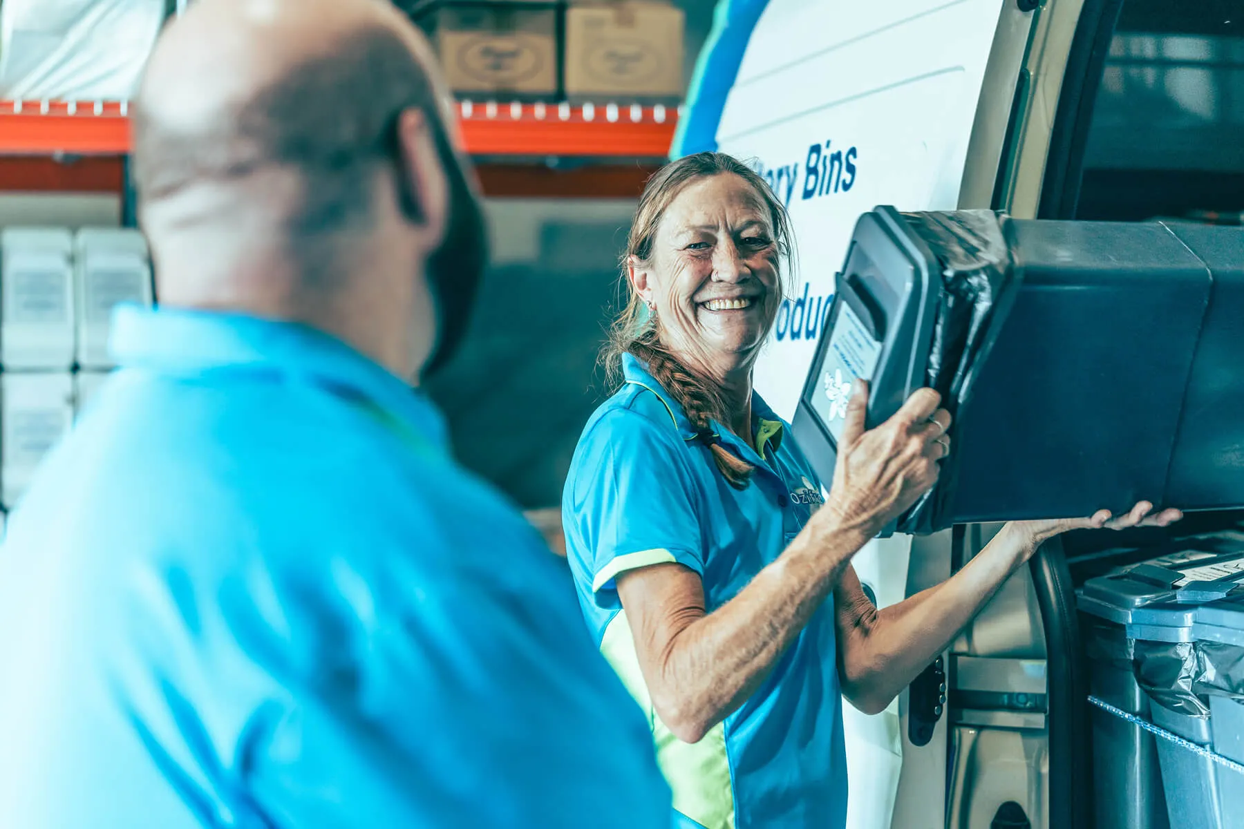 Smiling woman in blue uniform holding a black bin while interacting with a man in blue shirt in a warehouse setting.