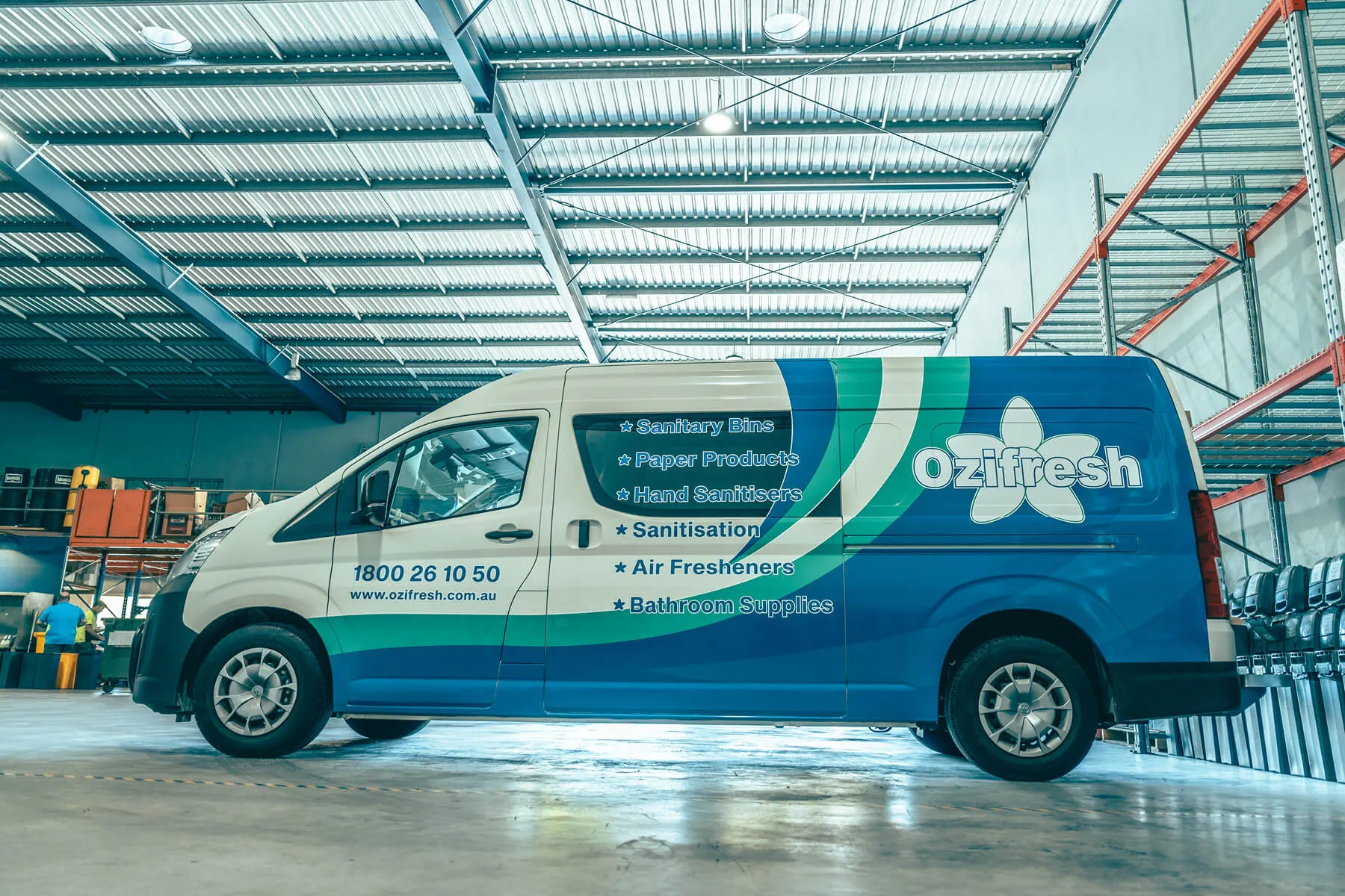Blue and white Ozifresh delivery van parked inside a warehouse with signage listing sanitary bins, paper products, hand sanitisers, sanitisation, air fresheners, and bathroom supplies.