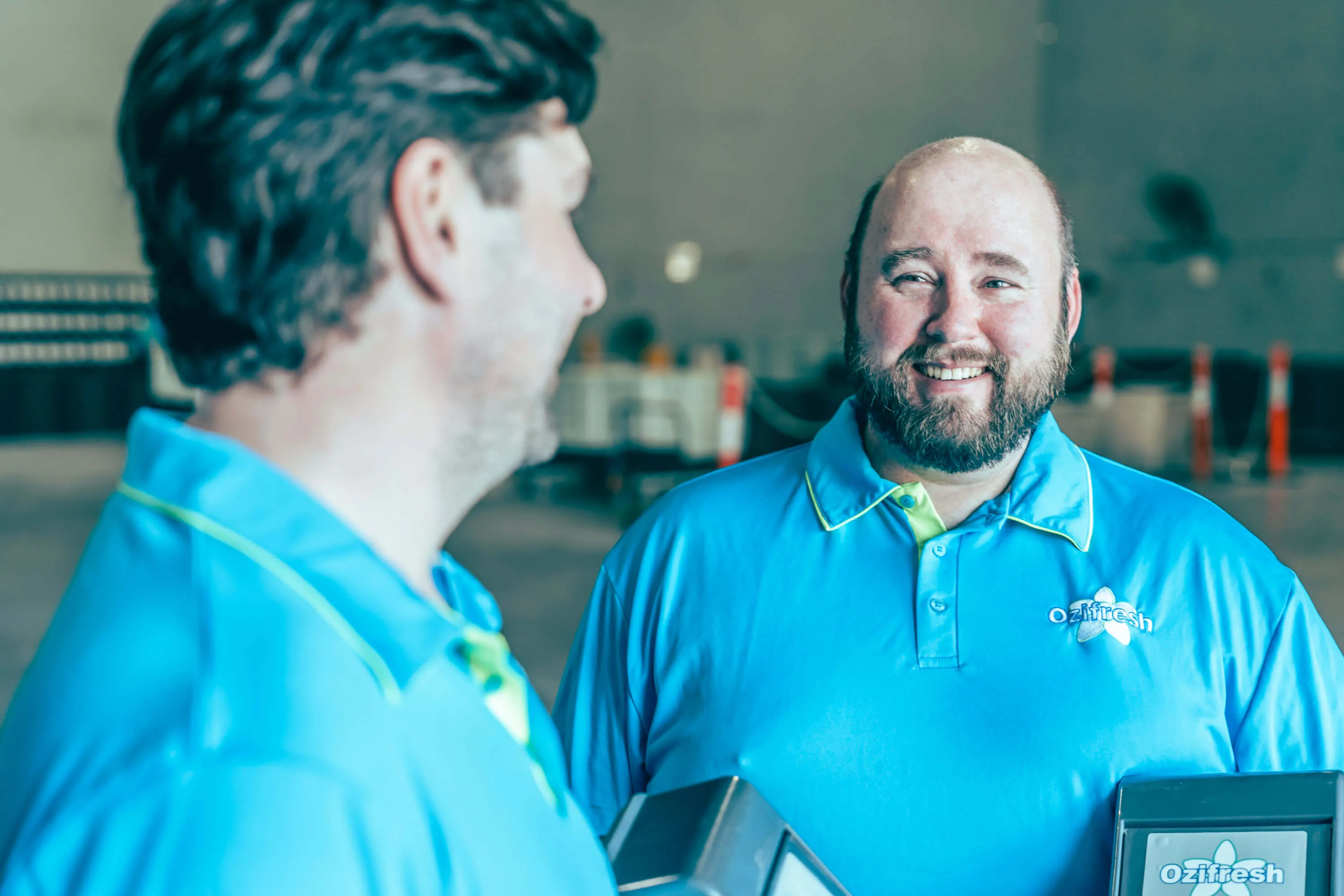 Two men in matching blue Ozifresh polo shirts smiling and talking in a warehouse-like setting.
