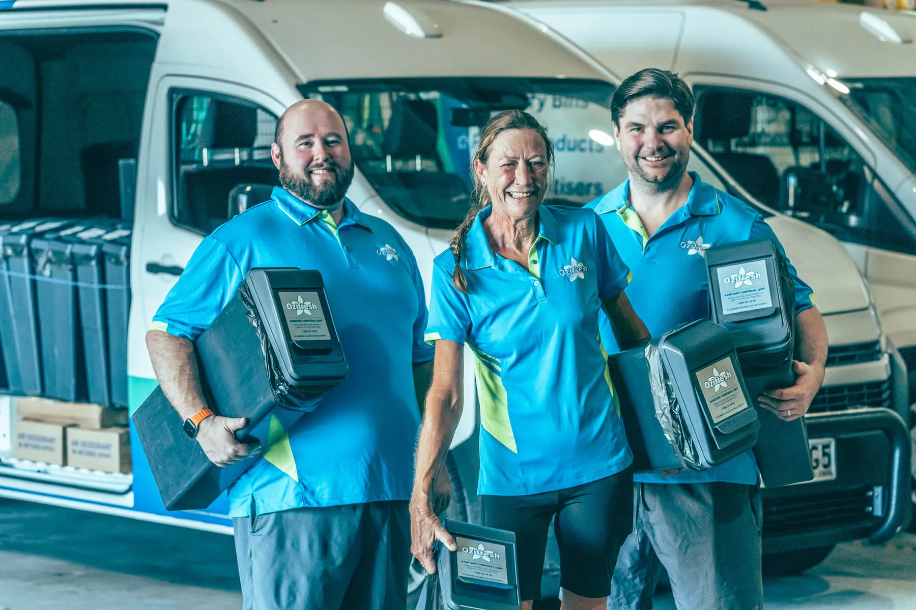 Three sanitation workers wearing matching blue uniforms standing in front of a white van, holding Ozifresh sanitation disposal units and smiling.