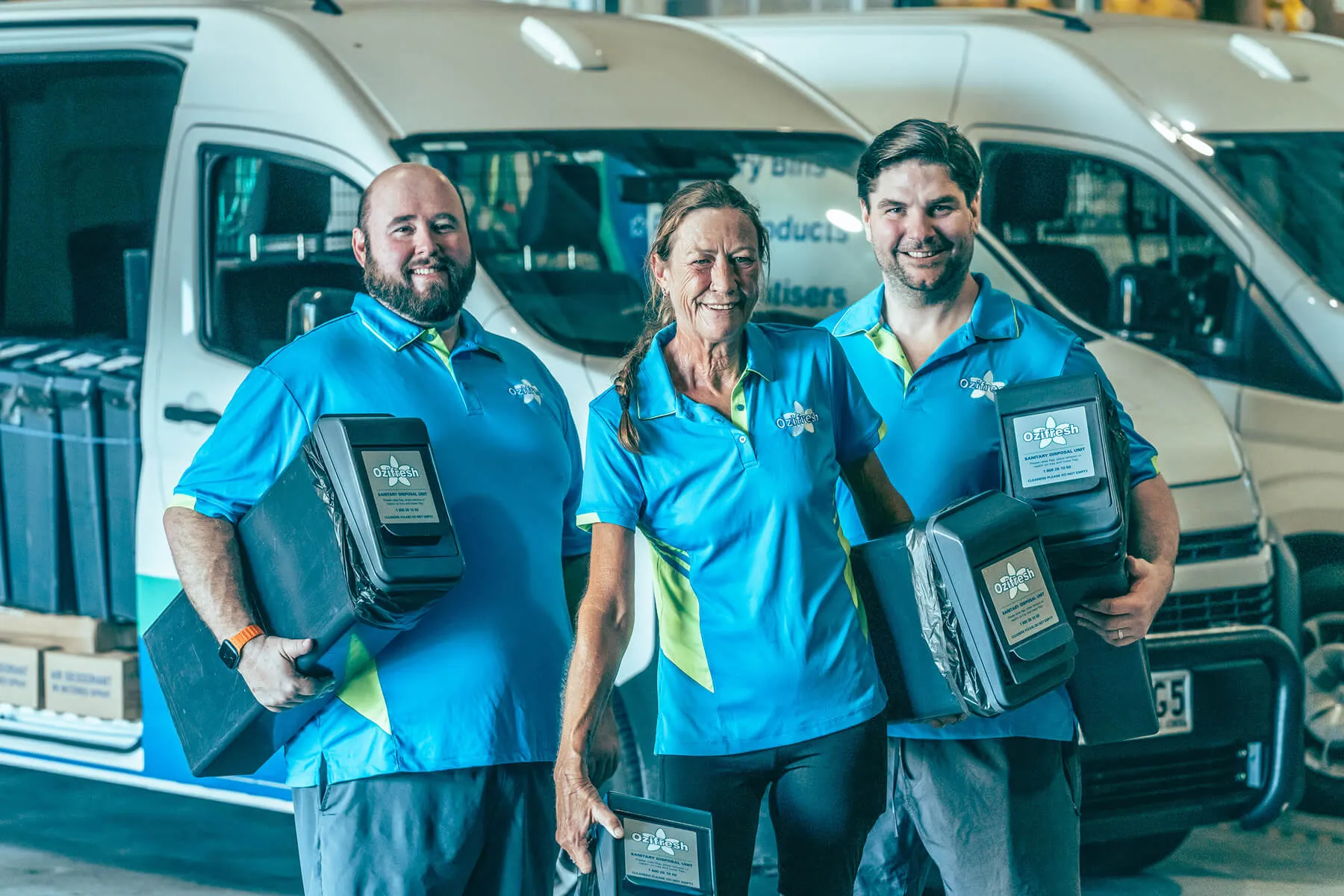 Three smiling workers in blue and green uniforms holding sanitary disposal units in front of white company vans.