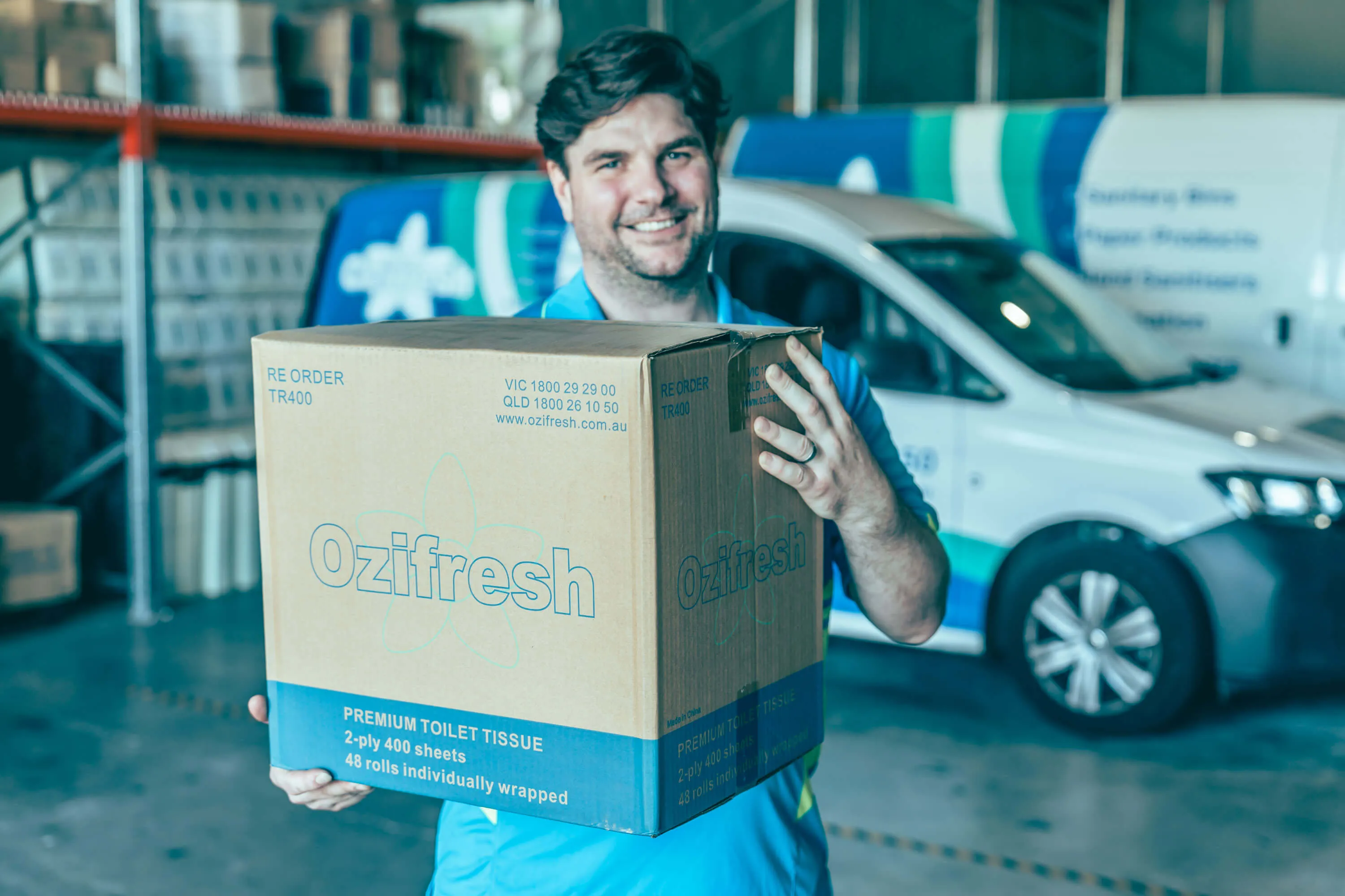 Smiling man holding a large box of Ozifresh premium toilet tissue in a warehouse with a company van in the background.