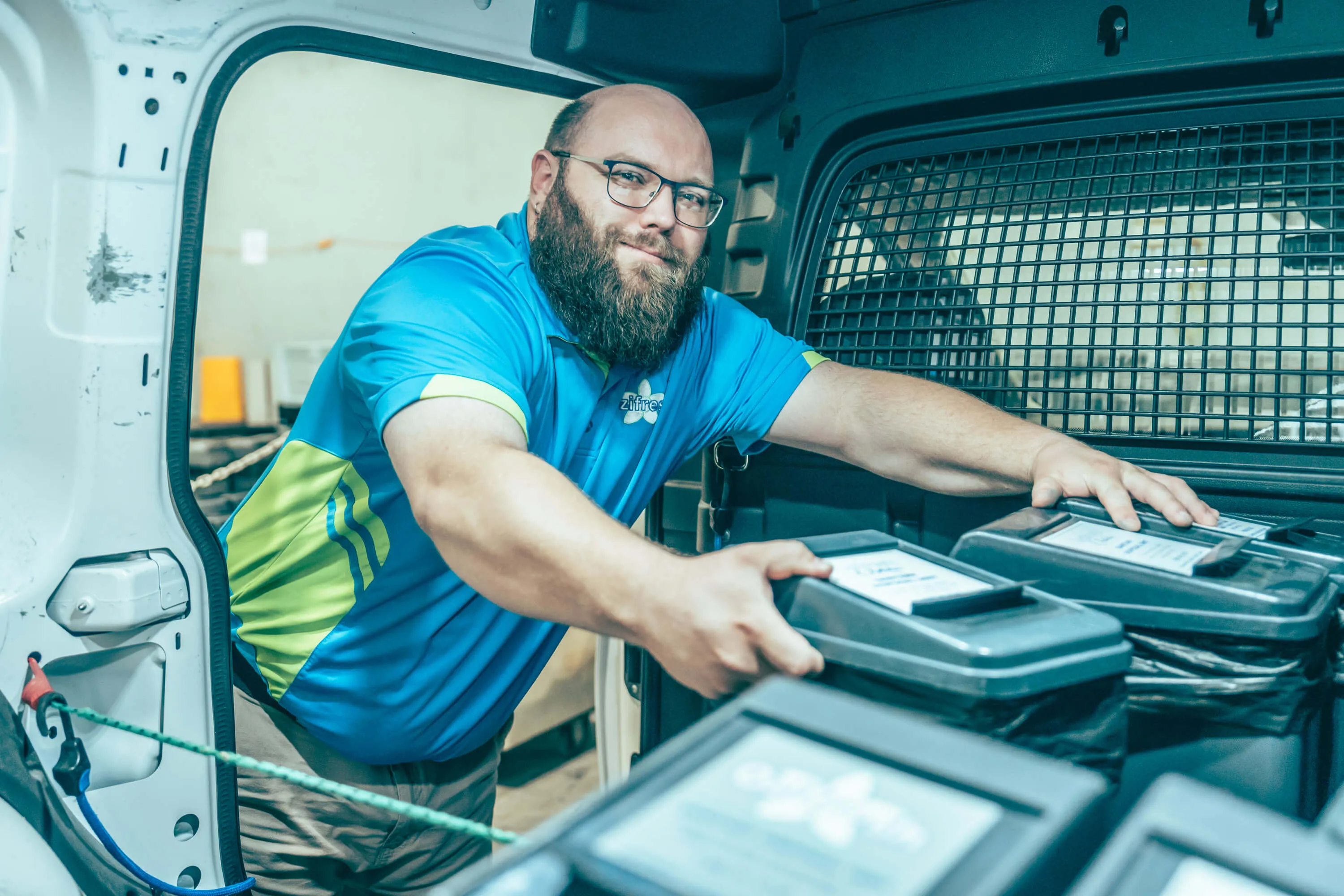Bearded man wearing glasses and a blue polo shirt loading labeled bins into the back of a van.
