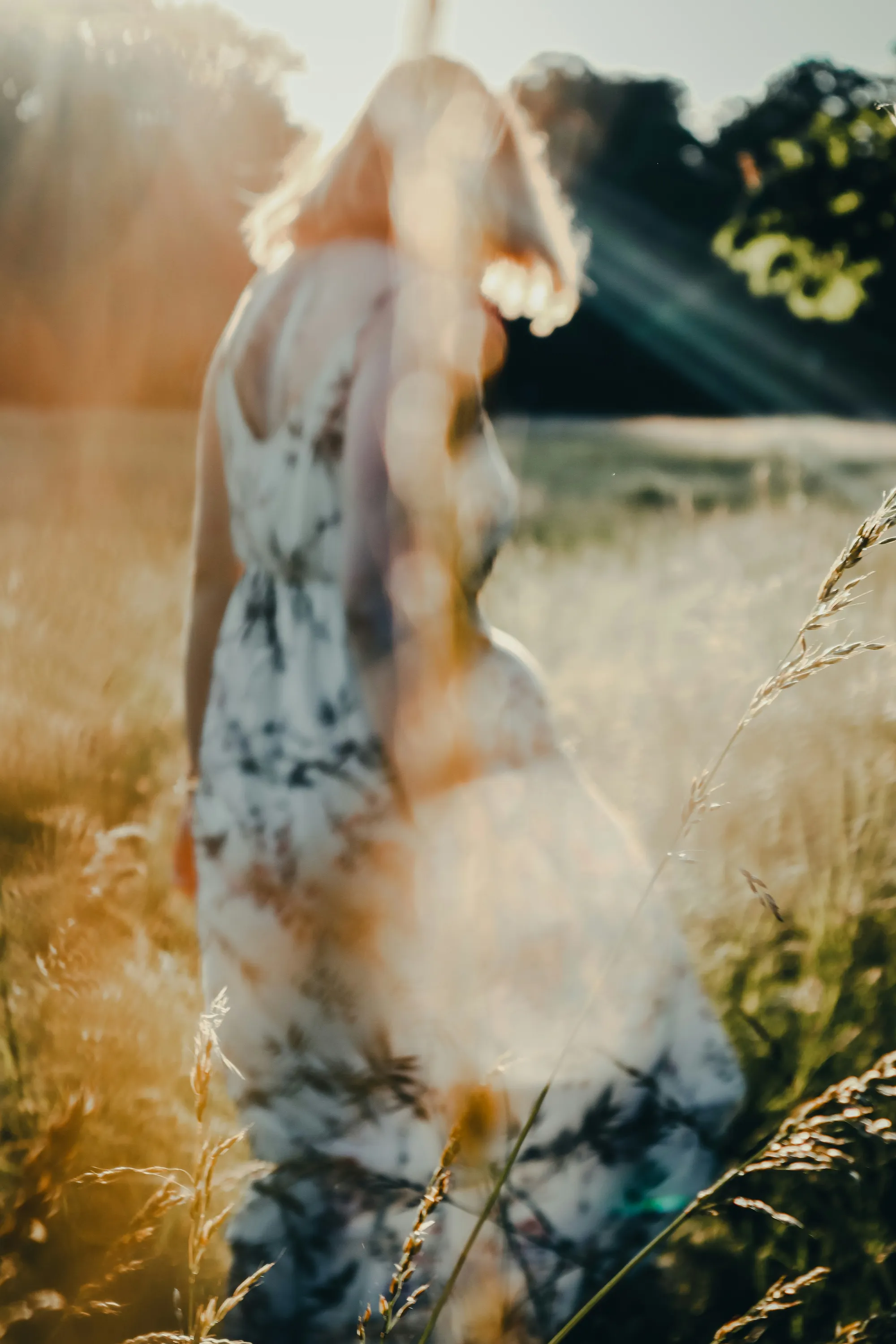 Woman in a floral dress walking through a sunlit field with tall grass and lens flare.