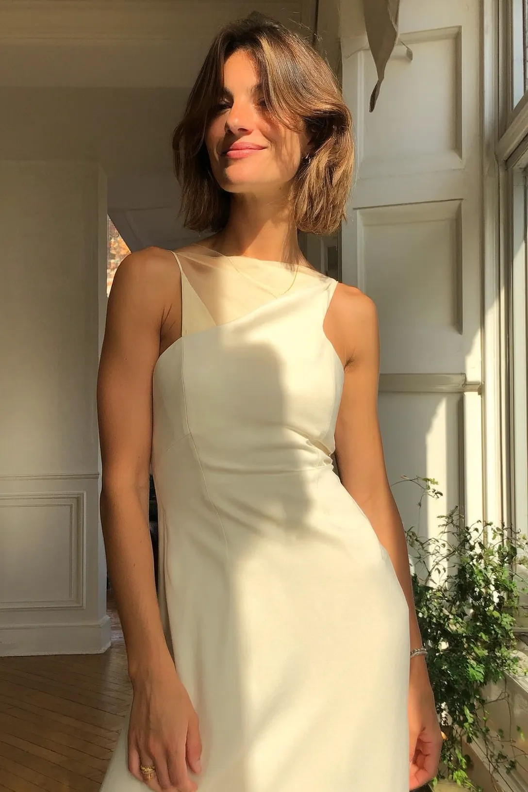 Woman with short brown hair wearing a sleeveless white dress standing near a window with sunlight and plants.