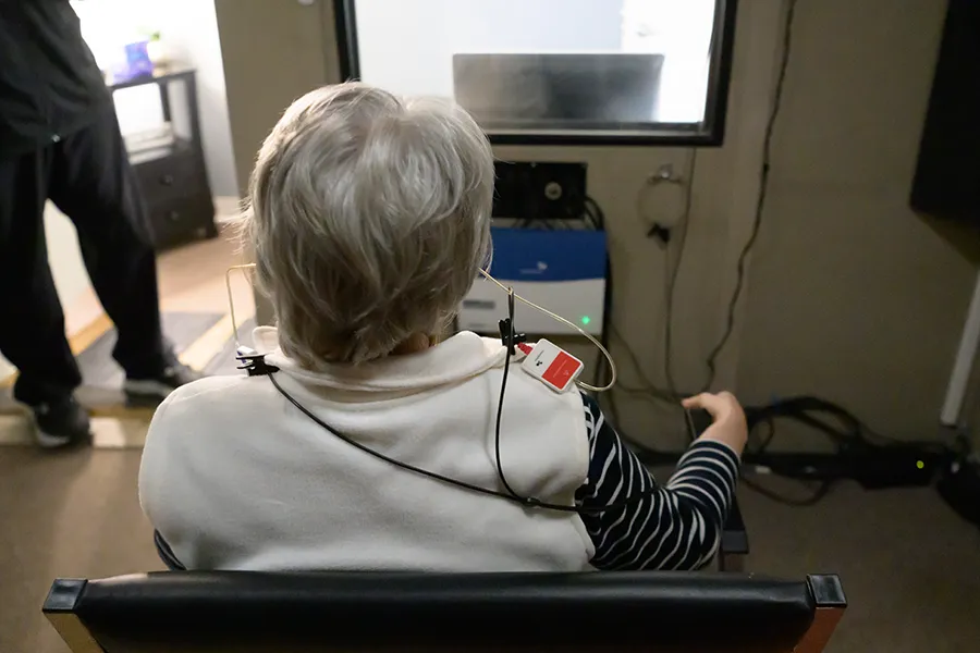 Person with gray hair wearing a hearing test device seated facing a machine in a clinic room.