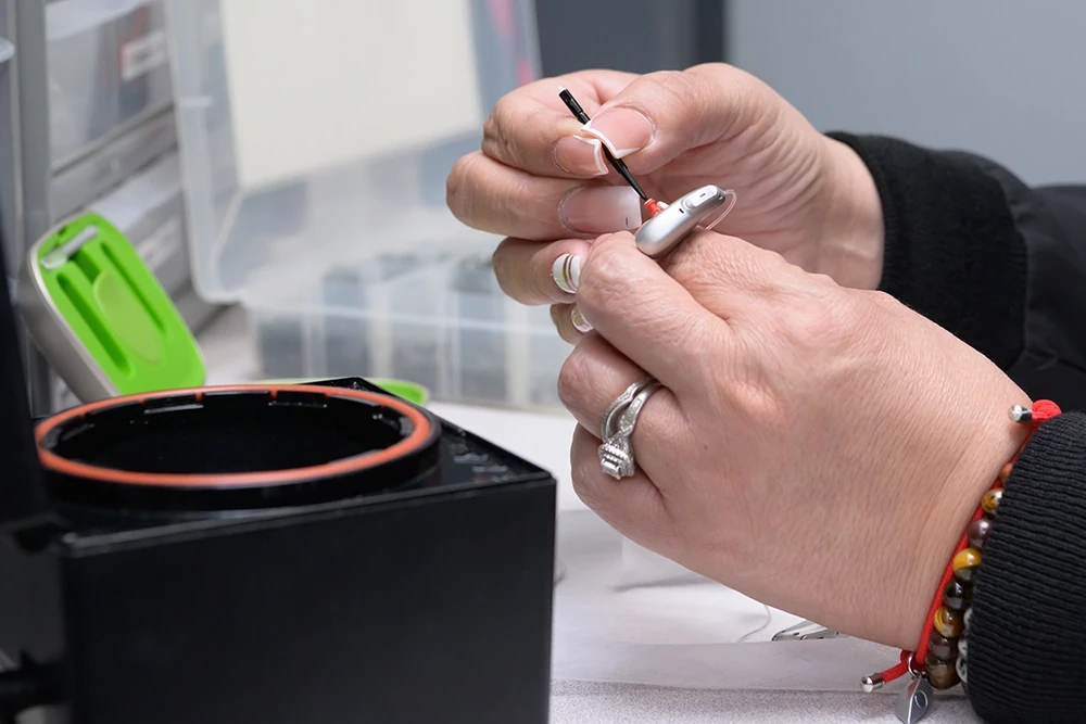 Hands holding and cleaning a small hearing aid device with a specialized tool on a work surface.