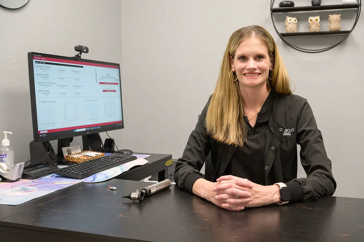 Female audiologist smiling and sitting at a desk with an otoscope and hearing aid on it, a computer with audiology software in the background.