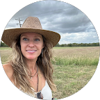 Smiling woman with long hair wearing a straw hat and necklace standing in a grassy field under a cloudy sky.