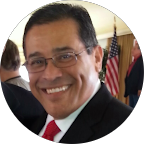Smiling man wearing glasses and a suit with a red tie, an American flag in the background.