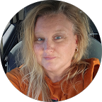 Woman with blonde hair and blue eyes wearing an orange top sitting inside a car.