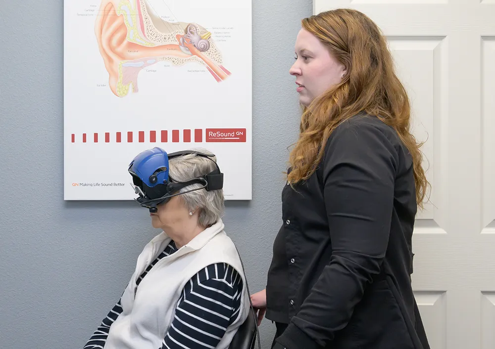 Older woman wearing an audiometric testing headset sitting in a chair with a technician standing beside her.
