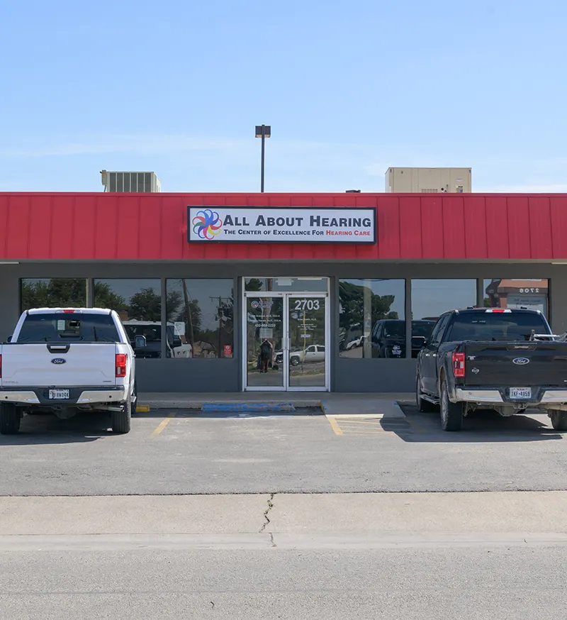 Front view of All About Hearing clinic with red awning and two parked Ford trucks in front.