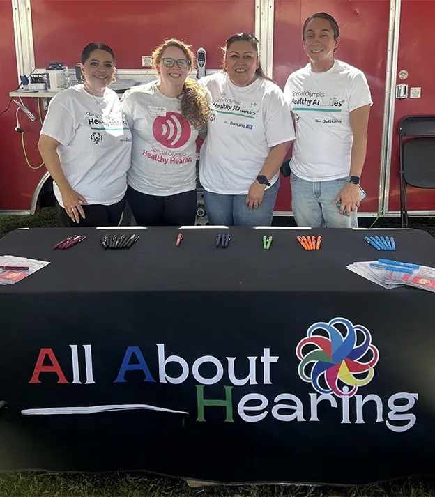 Person standing behind a booth table covered with informational pamphlets, hearing aids, and a rifle prop, with a black backdrop and a sign reading 'All About Hearing'.