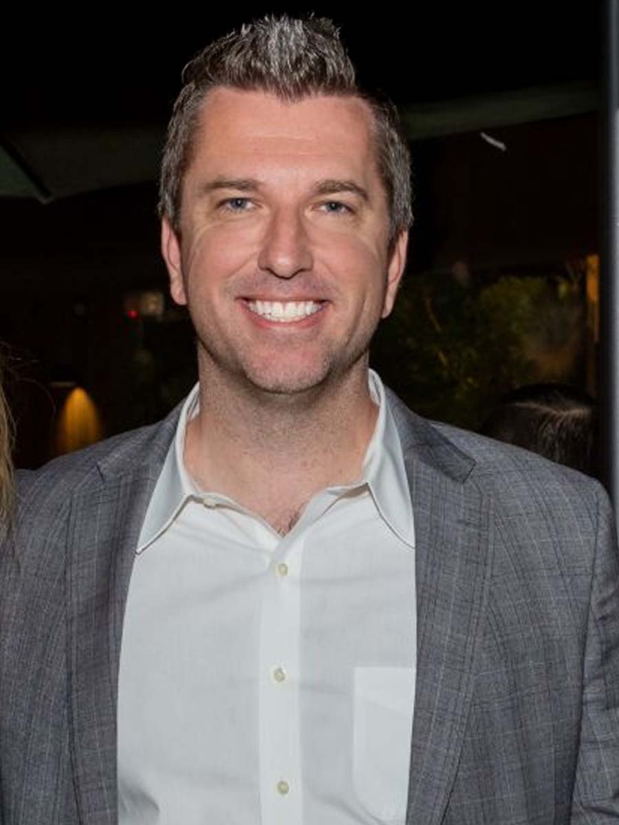 Smiling man with brown grey hair wearing a white buttoned blouse and beige pants standing against a two-tone gray wall.