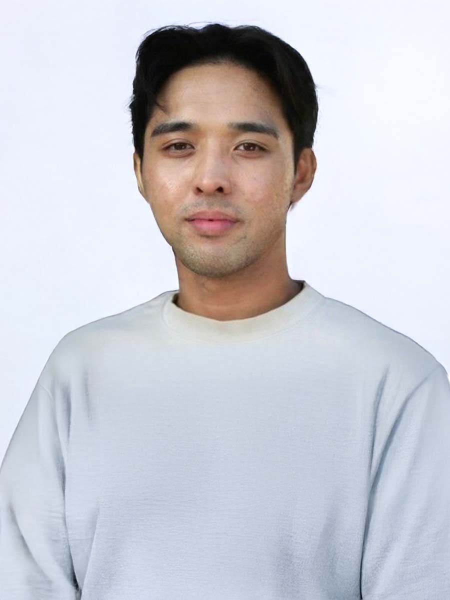 Young man smiling, wearing a white t-shirt and a wristwatch, standing outdoors with blurred greenery in the background.
