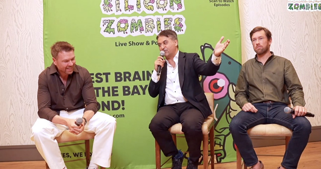 Three men sitting on chairs on a stage, one man in the middle speaking into a microphone with a green 'Silicon Zombies' live show banner behind them.