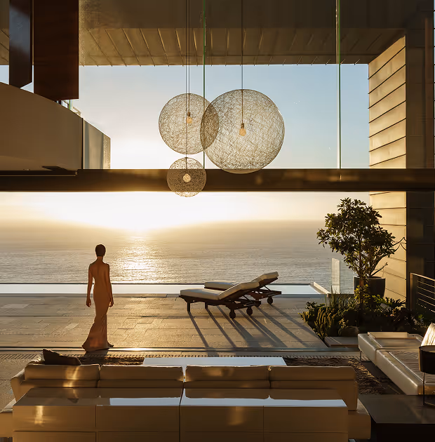 Woman in a long dress standing on a terrace overlooking a calm ocean at sunset, with lounge chairs and modern hanging light fixtures.