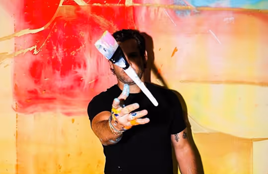 Man with paint-covered hands wearing a black shirt throwing a paintbrush in front of a colorful abstract background.
