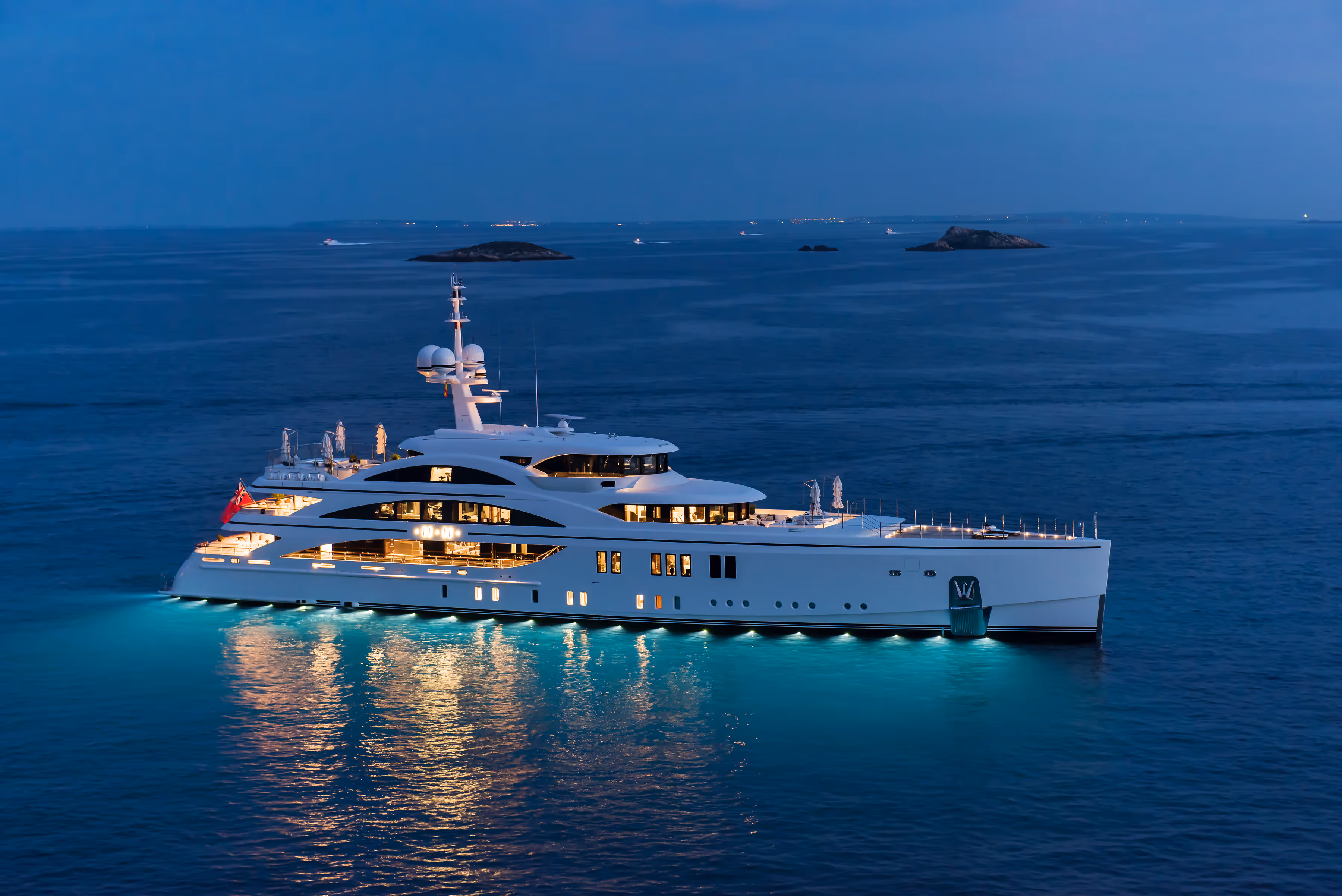 Luxurious white yacht illuminated with lights floating on calm ocean water at dusk with small rocky islands in the distance.