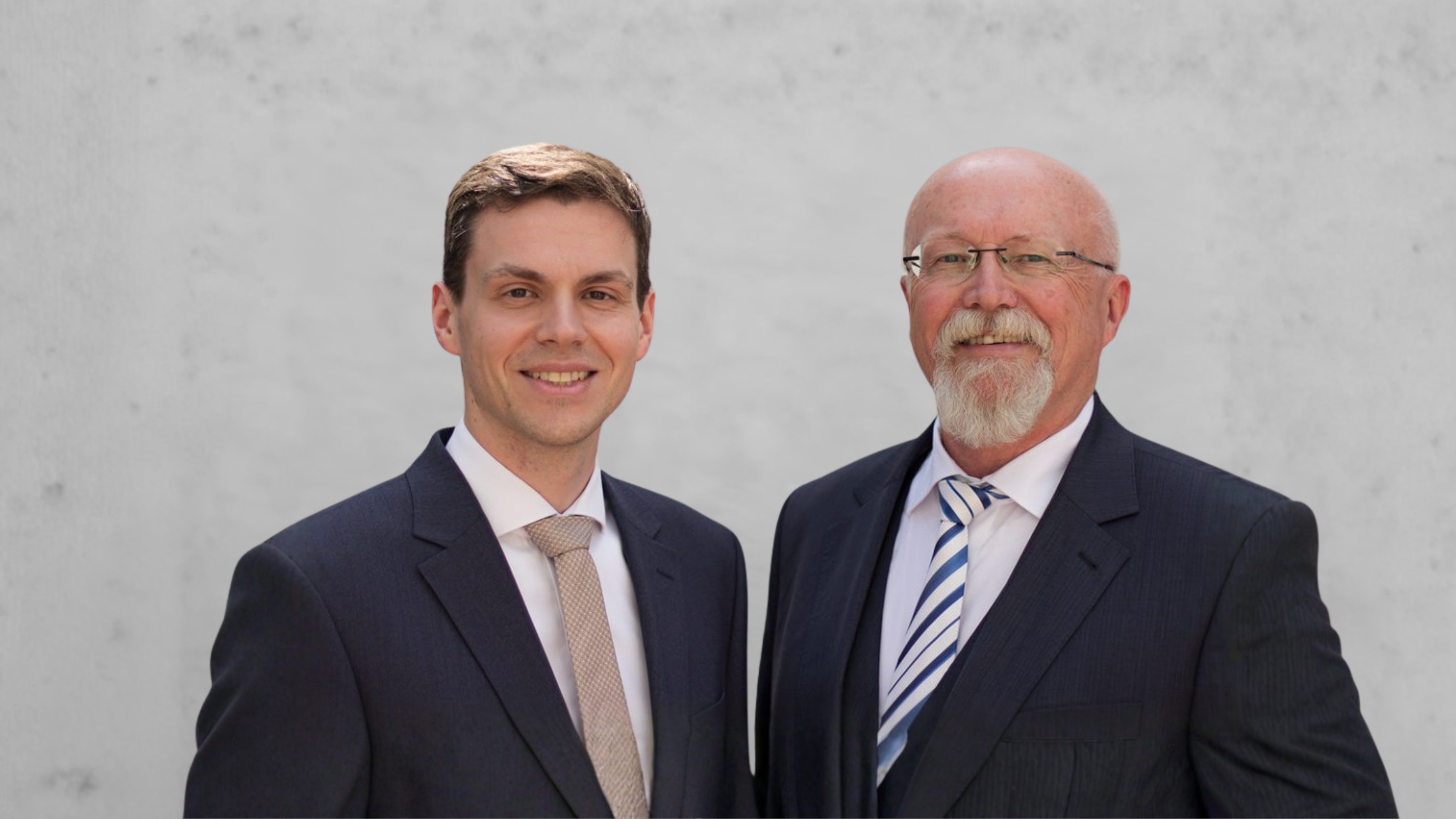 Zwei lächelnde Männer in Anzügen vor einem grauen Hintergrund, der jüngere trägt Krawatte in Beige, der ältere eine gestreifte Krawatte.