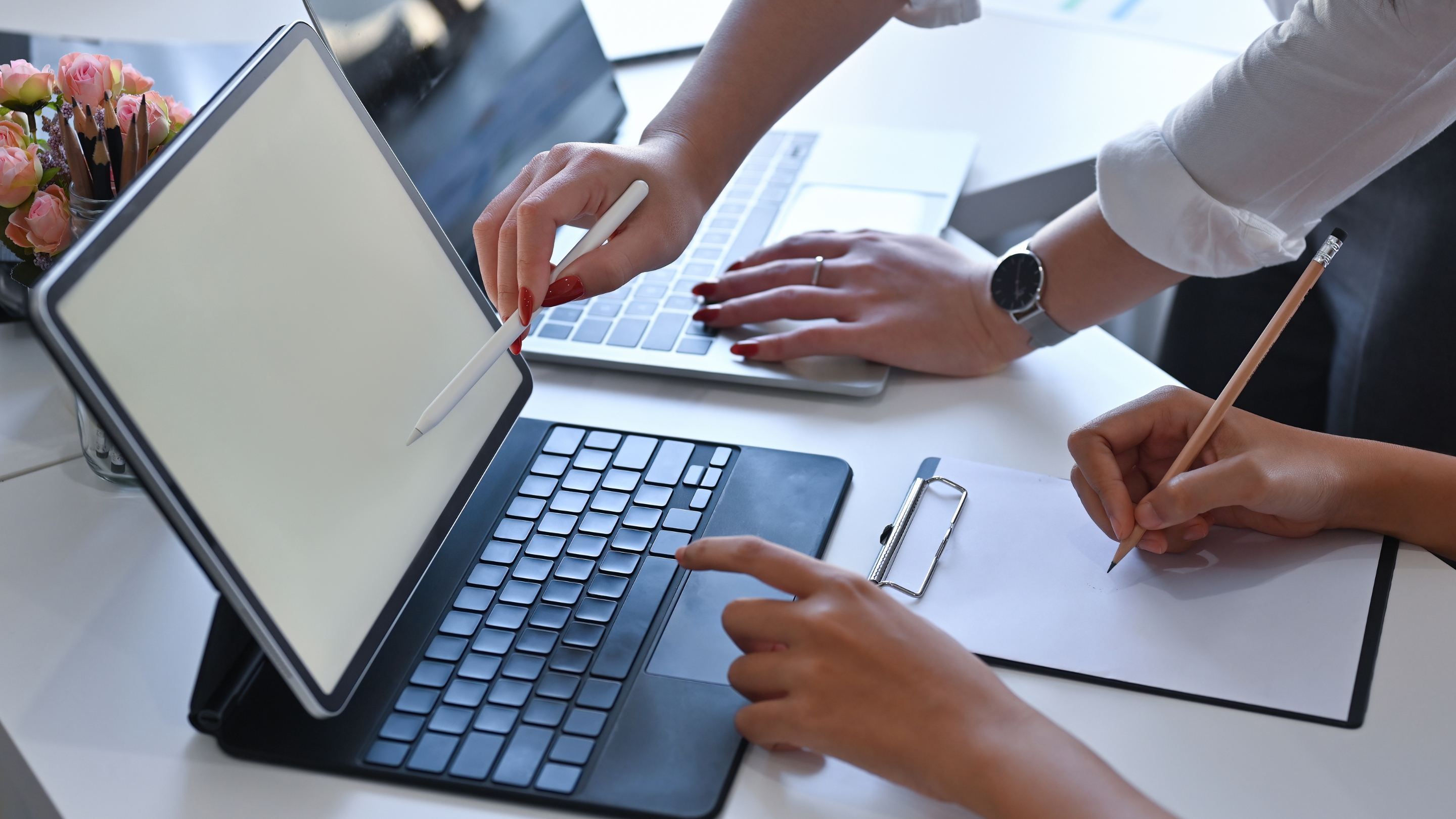 Zwei Personen arbeiten gemeinsam an einem Tablet mit Tastatur und einem Laptop, eine Person schreibt auf einem Klemmbrett mit einem Bleistift.