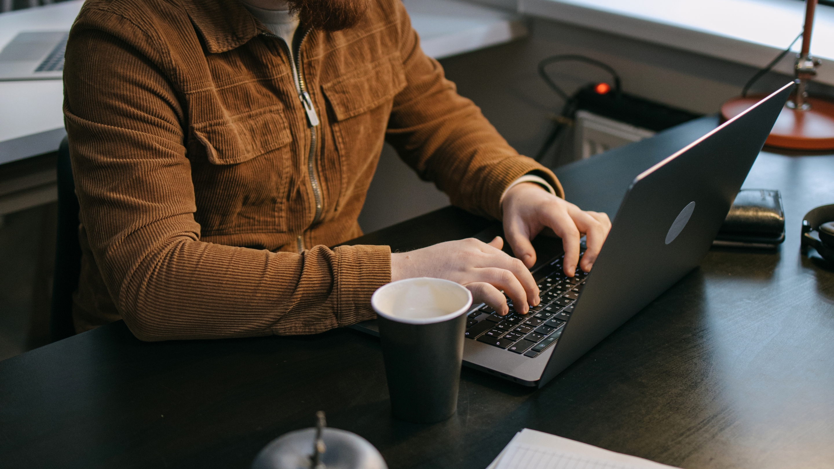 Person in braunem Cordjackett tippt auf einem Laptop an einem Schreibtisch mit einem Papierbecher.