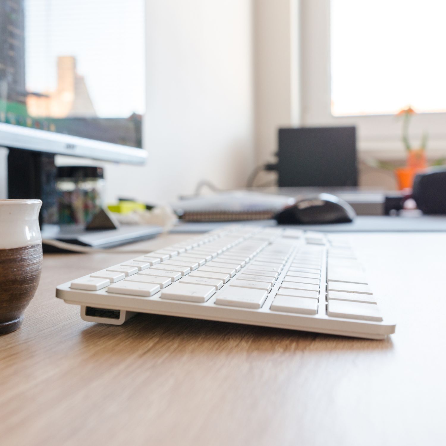 Nahaufnahme einer weißen Tastatur auf einem Schreibtisch mit Computermaus, Tasse und Monitor im Hintergrund.