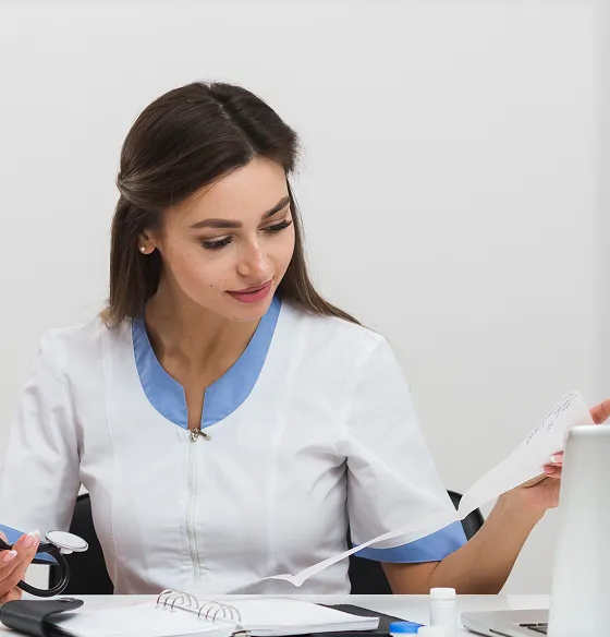 Female healthcare professional in white and blue uniform reviewing medical documents at desk.