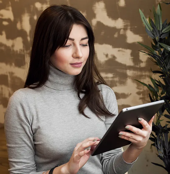Woman with long dark hair wearing a gray turtleneck sweater, using a tablet with a blurred brown and beige background and a plant beside her.