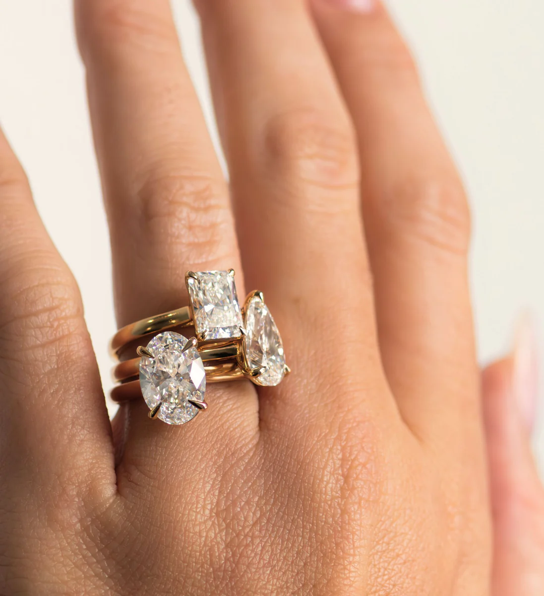 Hand wearing three gold rings with large oval, rectangular, and pear-shaped diamonds.