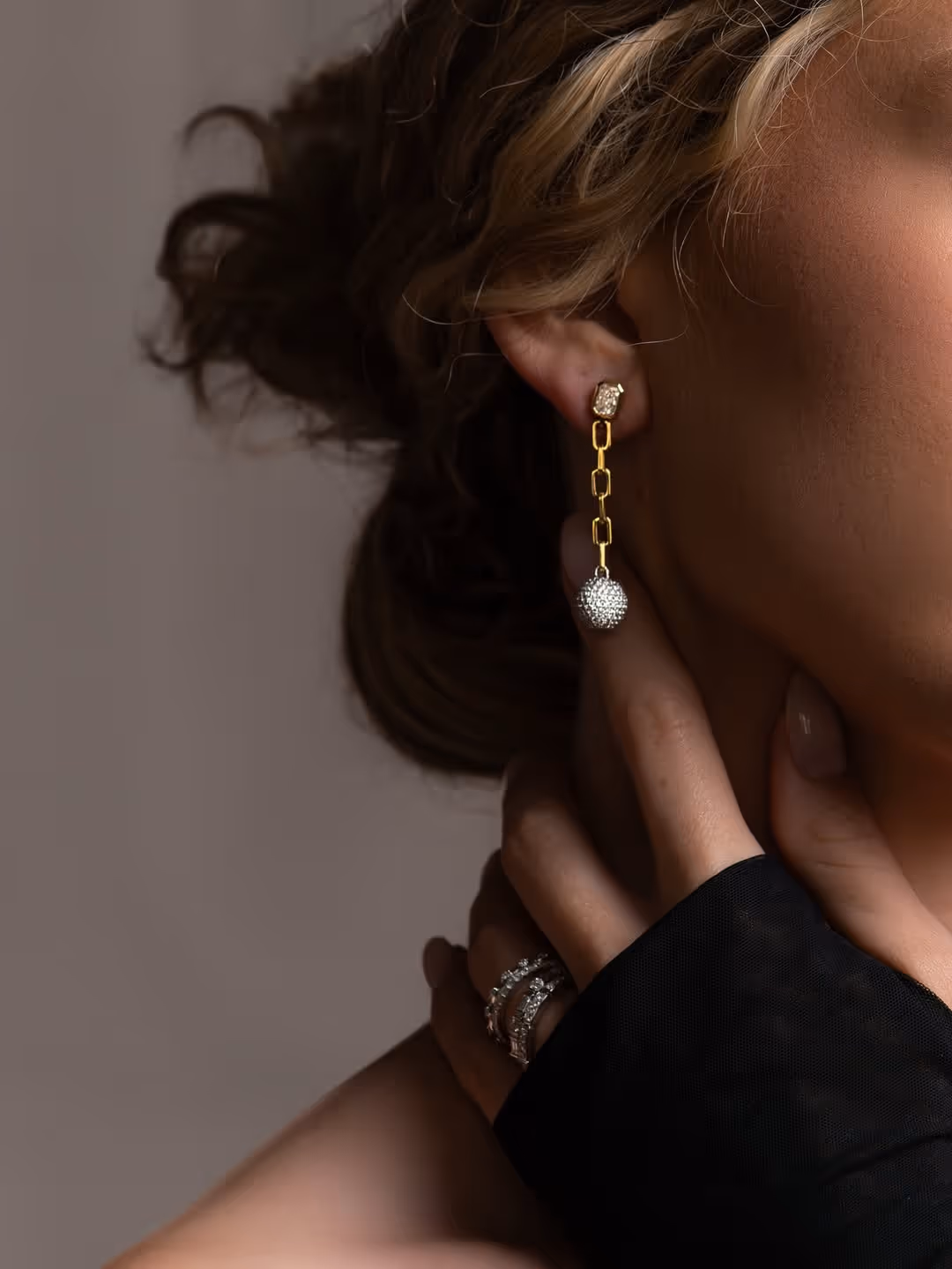 Close-up of a woman's ear wearing a gold chain earring with a small crystal stud and a sparkling spherical pendant, hand with diamond rings touching her neck.