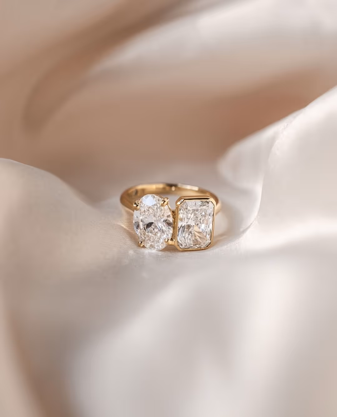 Gold ring featuring two large diamonds, one oval and one rectangular, displayed on soft cream fabric.