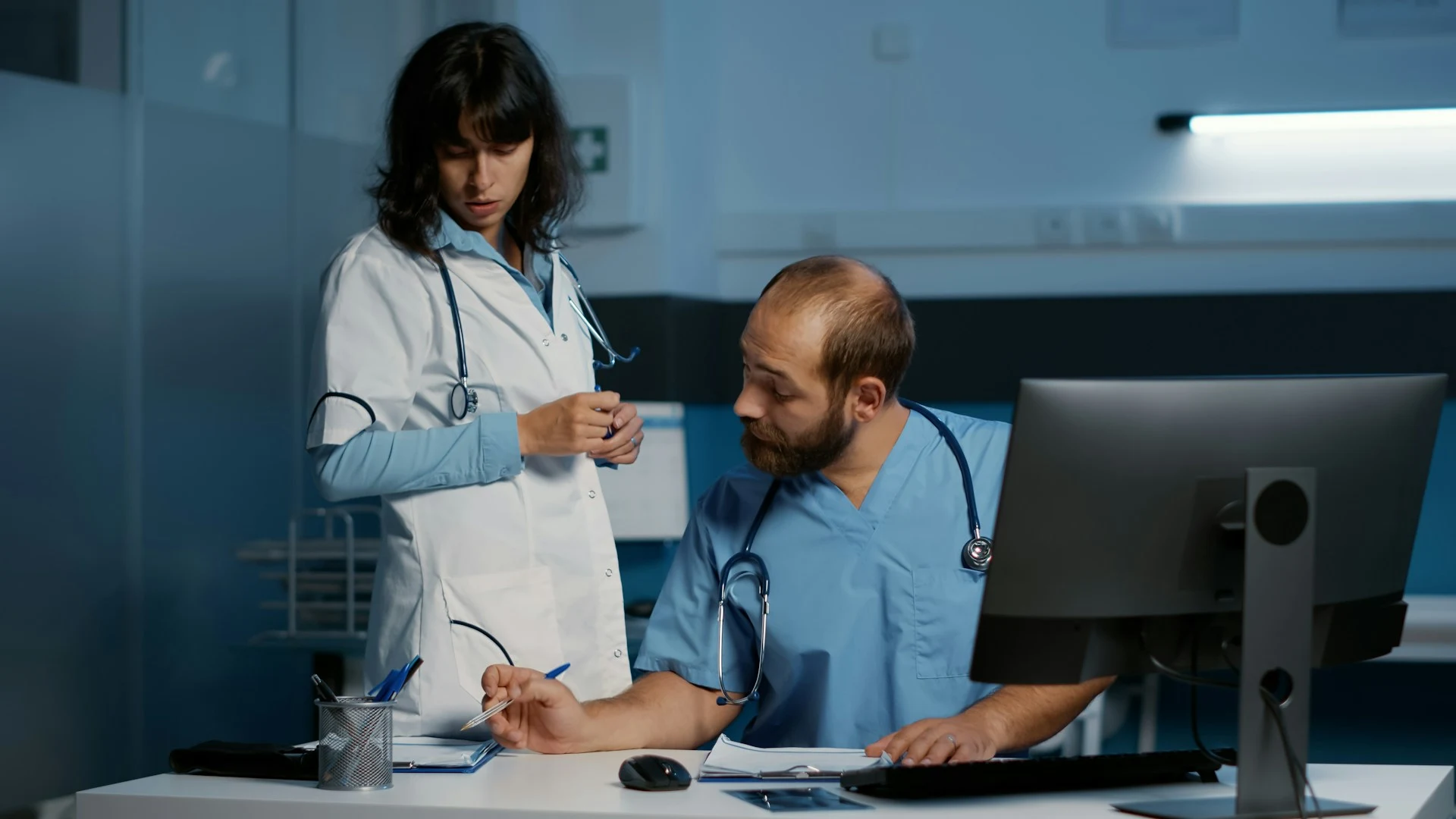 A male doctor seated at a desk with a computer discusses with a female doctor standing next to him in a hospital office.