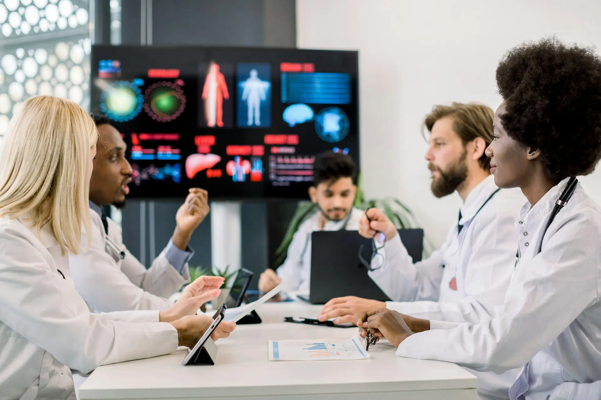 Group of diverse medical professionals in white coats discussing patient data in a meeting room with health charts displayed on a screen.