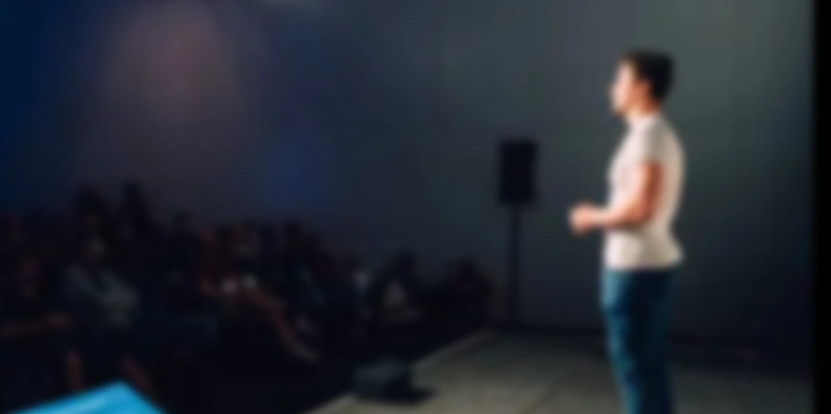 Blurry image of a person standing on a stage speaking to an audience seated in a dark room.