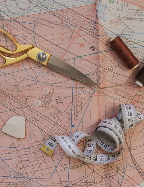 Sewing tools including gold-handled scissors, brown and black thread spools, white measuring tape, two white buttons, and fabric chalk on a sewing pattern sheet.