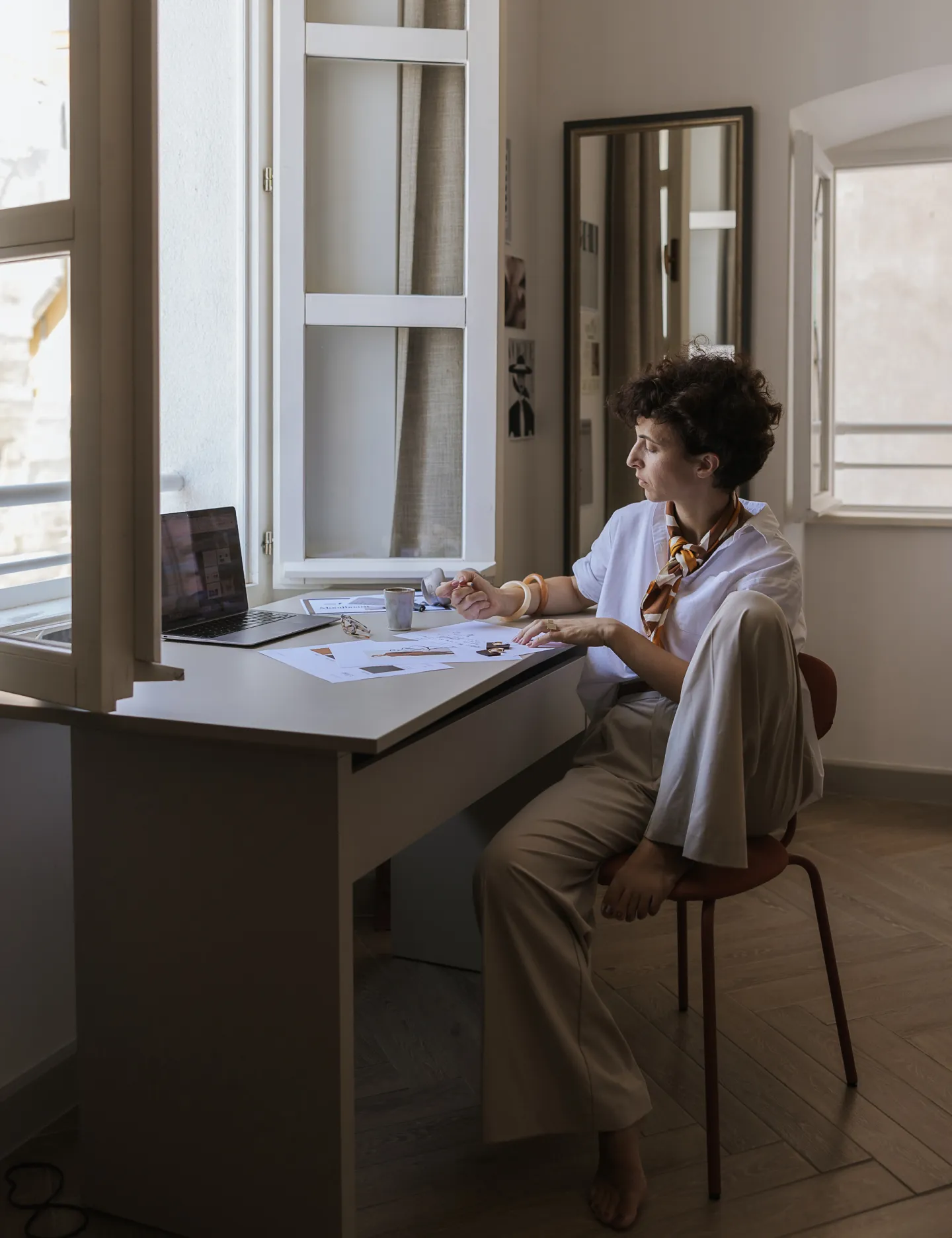 Woman sitting barefoot on a chair by a desk with papers and a laptop in a softly lit room with open windows.