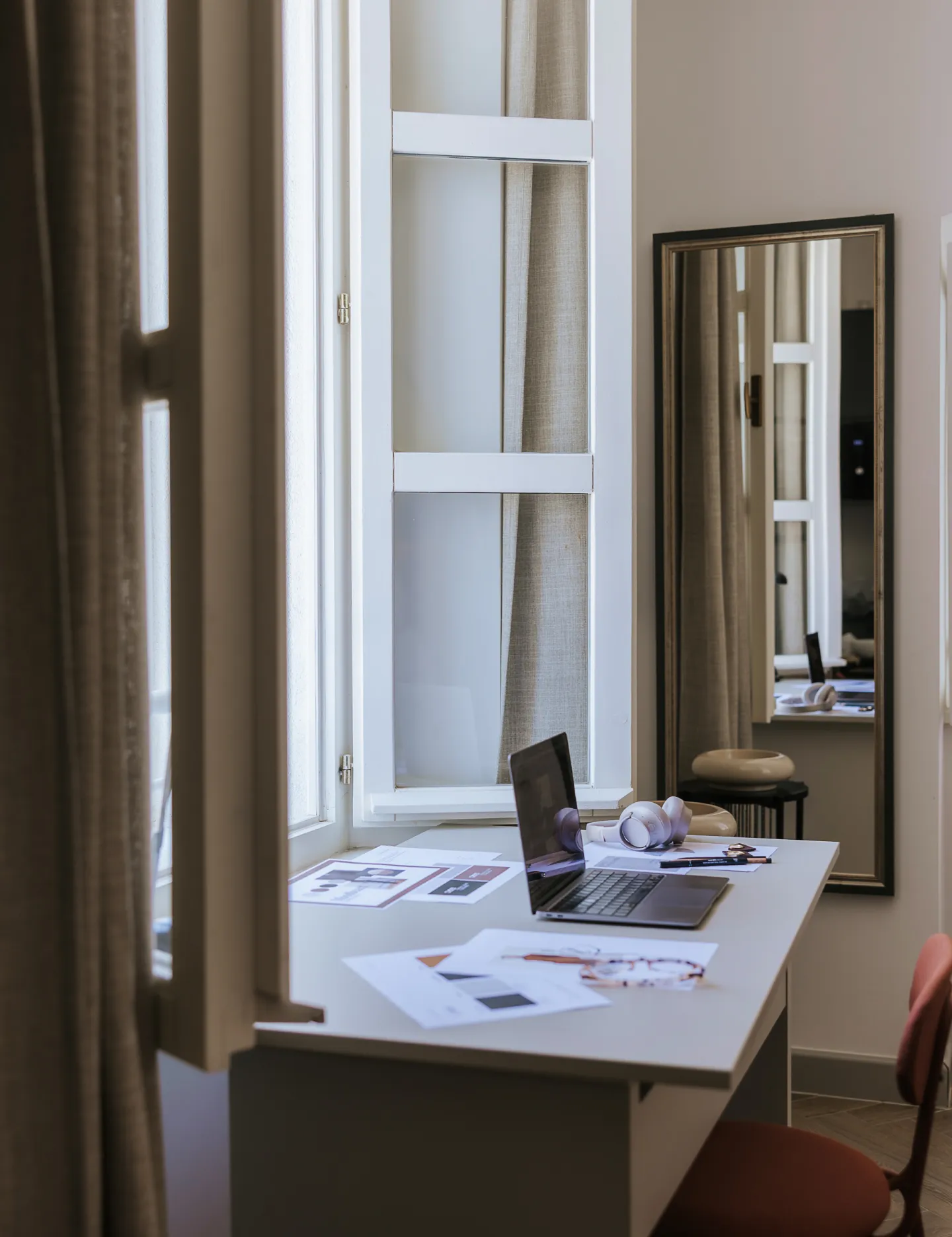 Minimalist workspace with a laptop, headphones, papers on a white desk near a window and a wall mirror reflecting the setup.