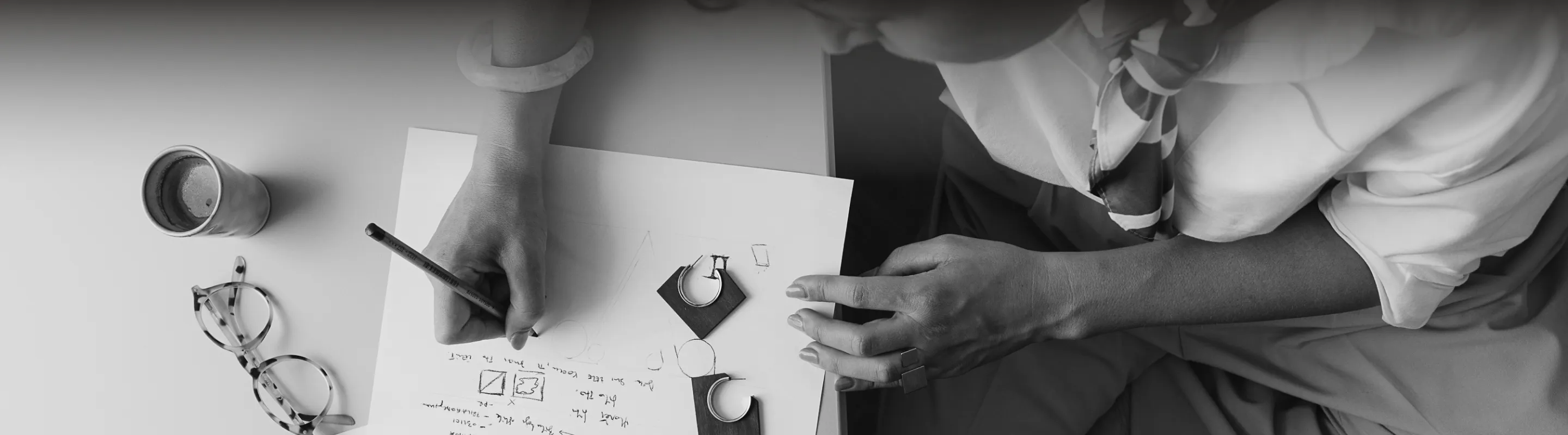 Person sketching jewelry designs on paper with a pencil, surrounded by eyeglasses and a cup on a table.