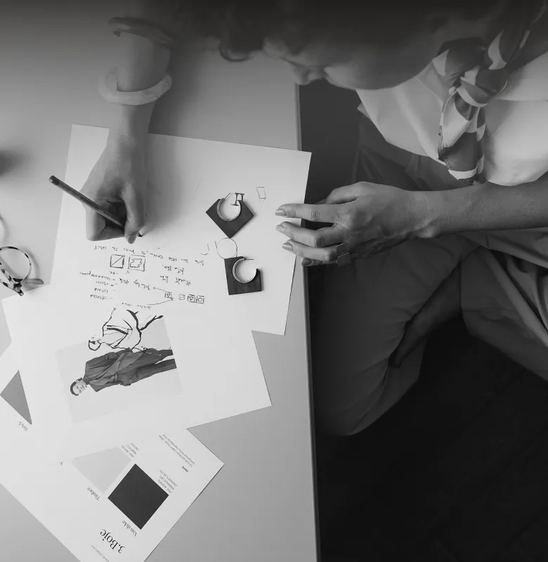 Person sketching fashion designs and notes on paper at a desk with earrings and fabric swatches.